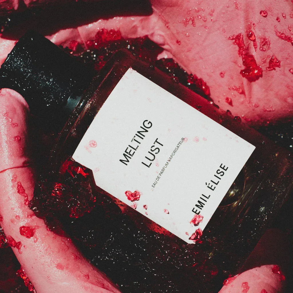 Flasche eines Parfums mit der Aufschrift "Melting Lust" von "Emil Élise", umgeben von roter Flüssigkeit und rosa Textur.