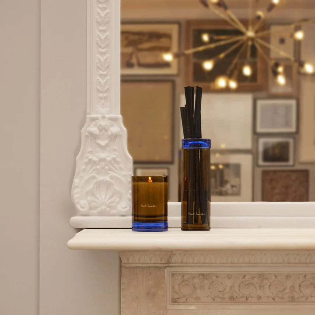 Der Storyteller Diffuser von Paul Smith zeigt einen braunen Glasbehälter mit blauen Akzenten und schwarze Duftstäbchen.