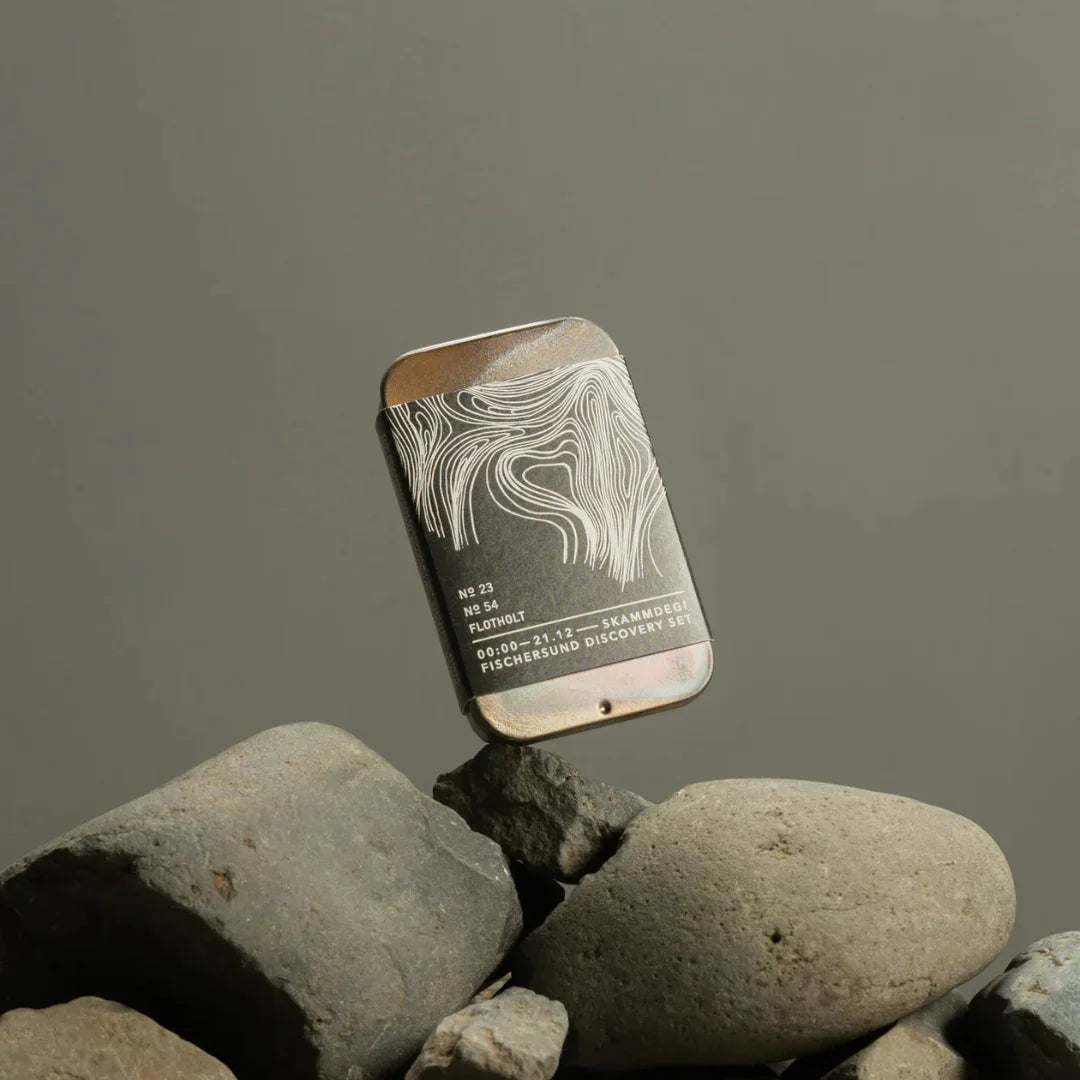 Metallische rechteckige Dose auf Steinen, verziert mit abstrakten Linien. Beschriftet mit "NR 23", "Fischersund", "Skammdegi Discovery Set".