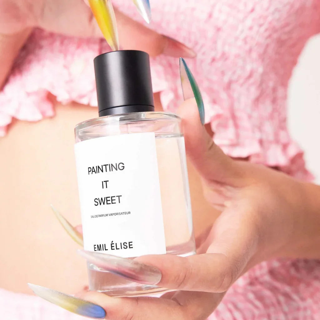 Frau mit langen, bunten Nägeln hält eine Flasche Parfüm mit der Aufschrift "Painting it Sweet" von Emil Élise in der Hand.