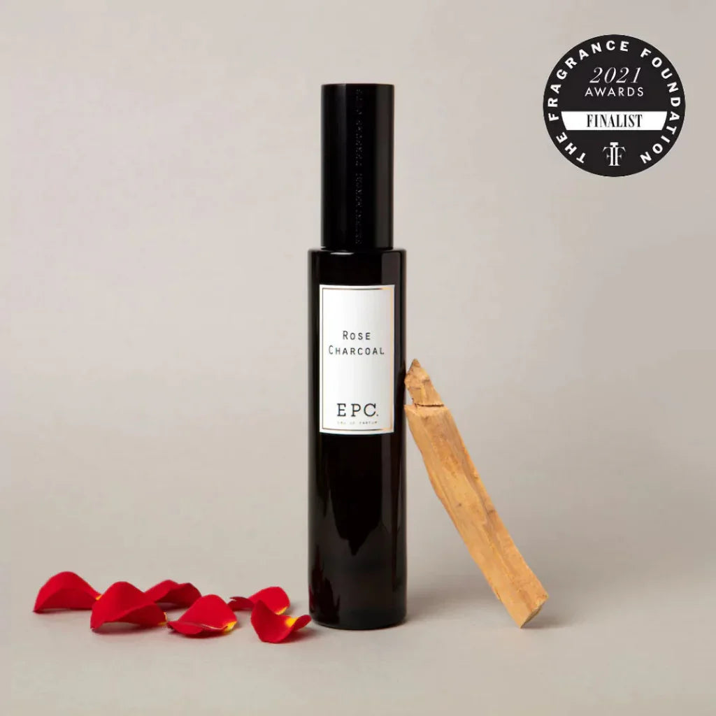 Dunkle Glasflasche mit schwarzem Verschluss und weißem Etikett, "Rose Charcoal" von Experimental Perfume Club, mit Rosenblättern und Holzstück.
