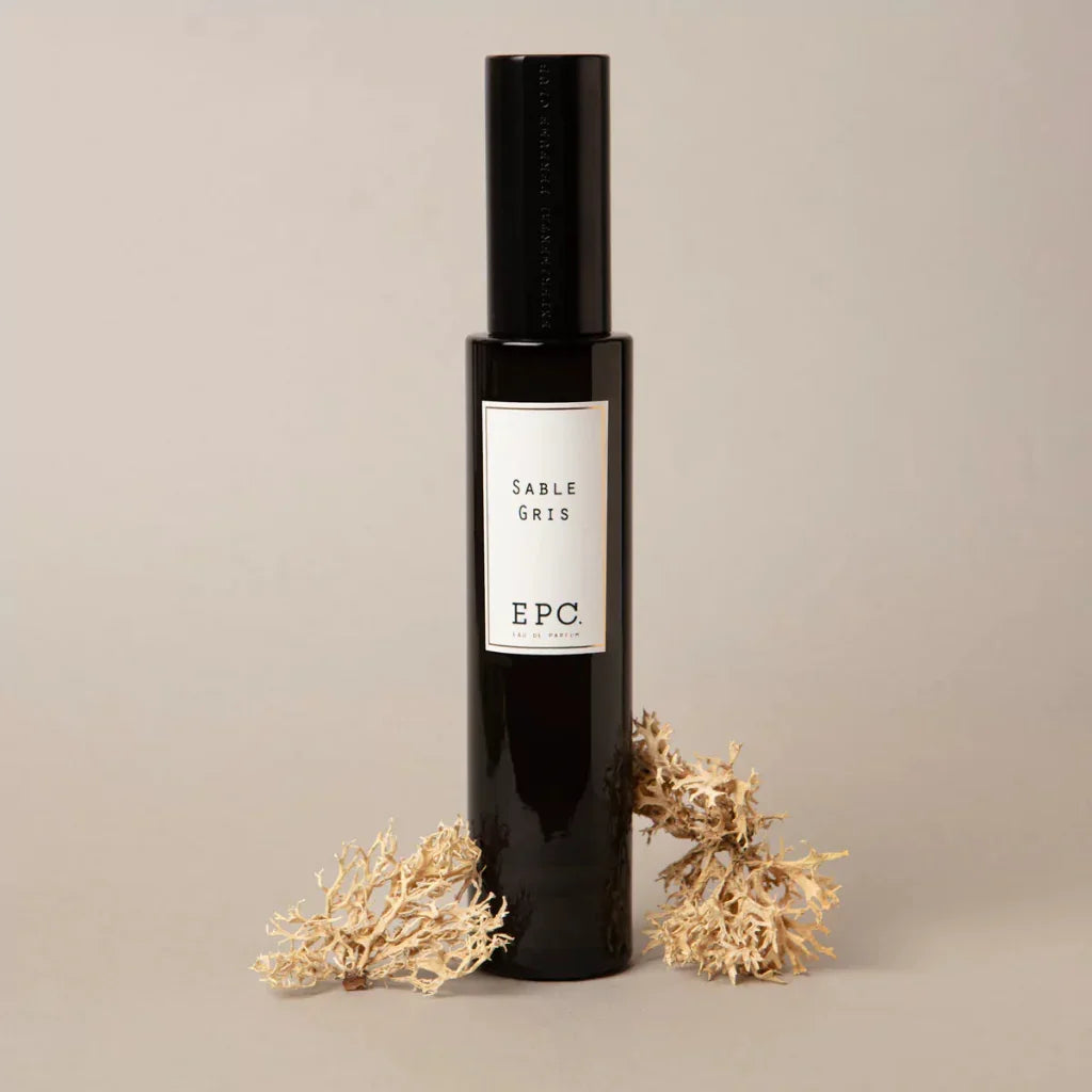 Dunkle, schlanke Parfumflasche mit weißem Etikett "Sable Gris" von Experimental Perfume Club, flankiert von getrockneten Pflanzen.