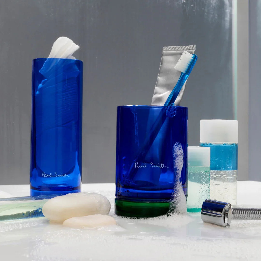 Das Bild zeigt blaue Glasbehälter mit der Aufschrift "Paul Smith", gefüllt mit Toilettenartikeln, neben Seifenspendern und Flüssigkeiten auf einem nassen Untergrund. Produktname: Early Bird Duftkerze, Anbieter: Paul Smith.