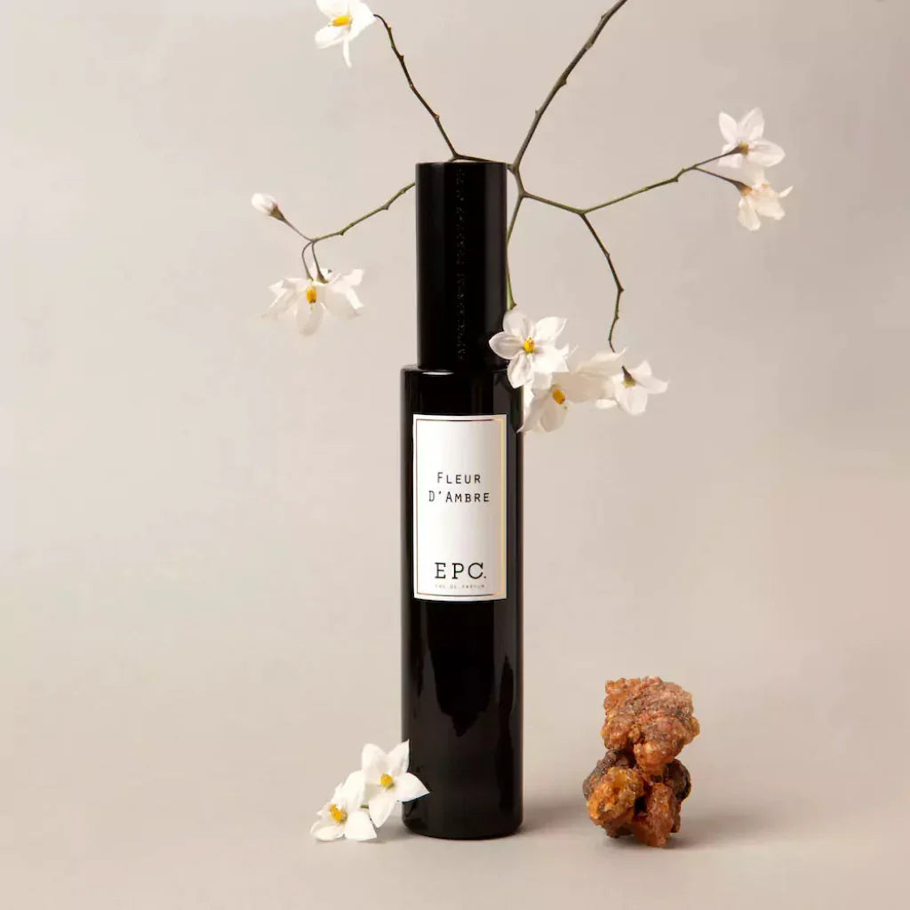 Schwarzer schlanker Flakon "Fleur d'Ambre" von Experimental Perfume Club mit weißen Blumen und einem Harzstück daneben.
