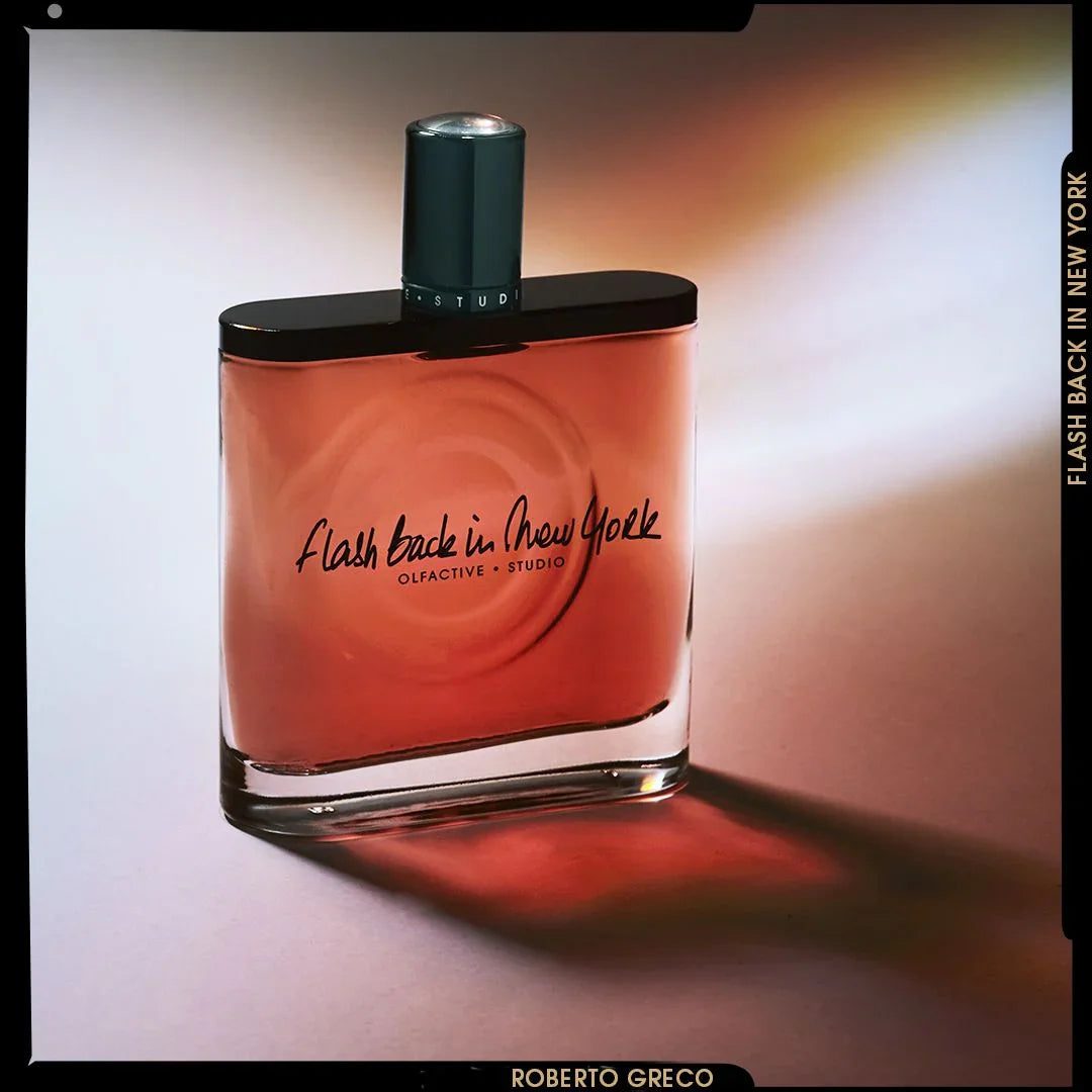Parfümflasche mit orangefarbener Flüssigkeit und schwarzem Sprühkopf, Text "Flash Back in New York" und "Olfactive Studio".