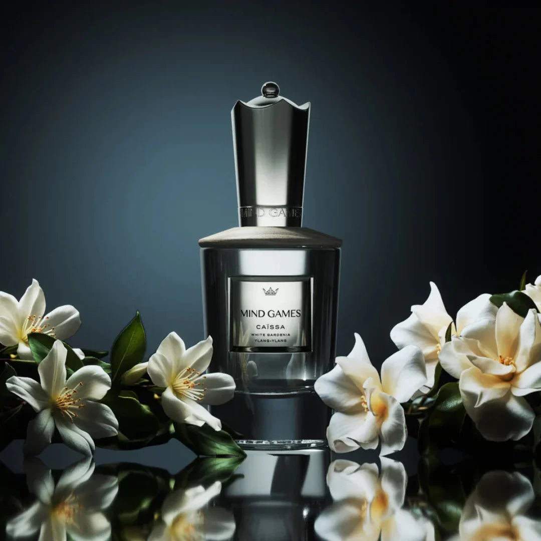 Flakon des Parfüms "Caïssa" von Mind Games, elegant gestaltet in silberner Farbe mit weiß-texturierten Akzenten, umgeben von weißen Blüten.