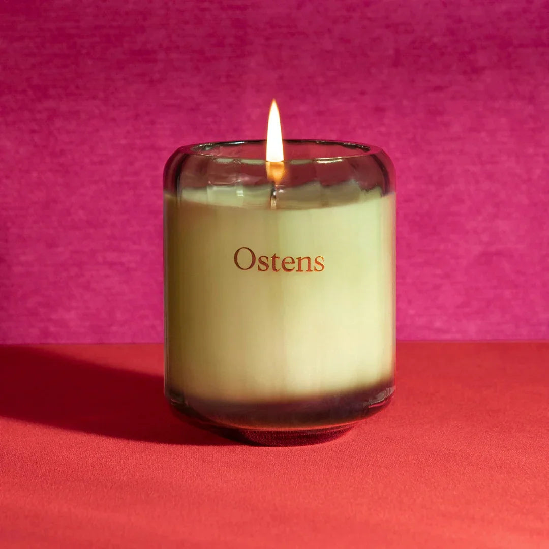 Illumination Rose Duftkerze von Ostens, eine brennende Kerze in einem transparenten Glas mit "Ostens"-Schriftzug.