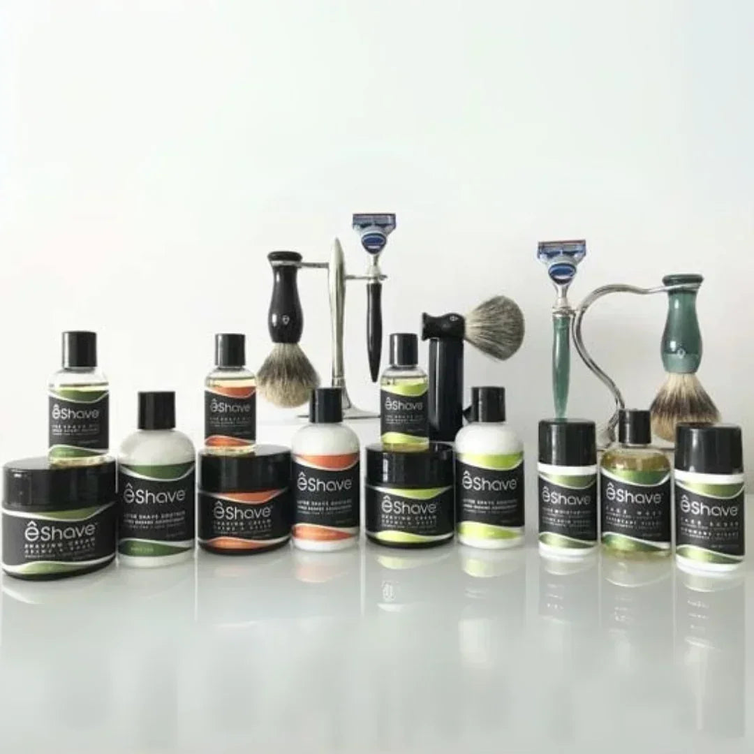 Produktauswahl von eShave, darunter "Waschlotion Weisser Tee" in runden Flaschen mit schwarzem Etikett. Pinsel und Rasierer im Hintergrund.