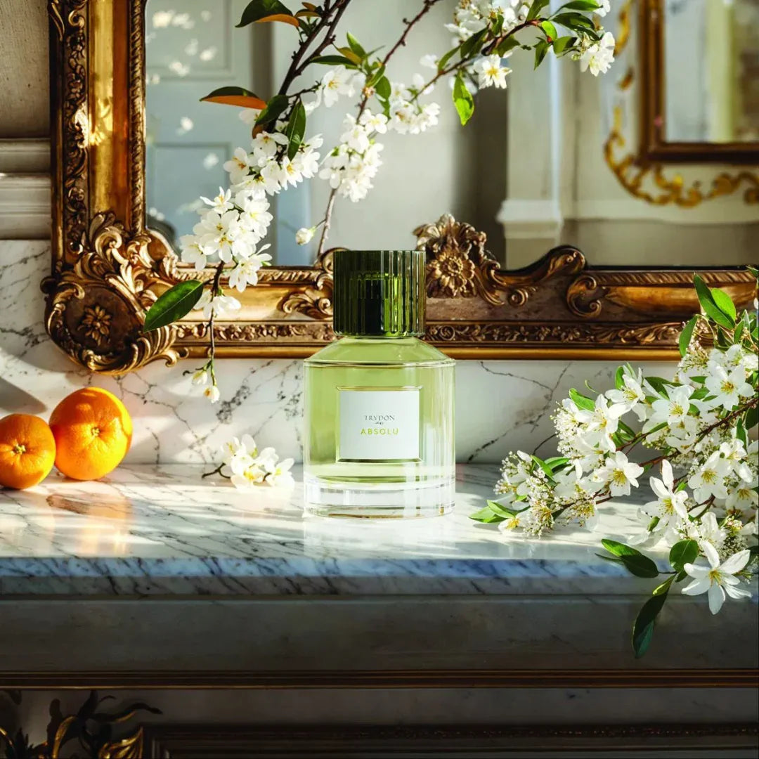 Duftflakon "ABSOLU" von Trudon, mit grünem Deckel und klarem Glas, umgeben von weißen Blüten und Orangen.
