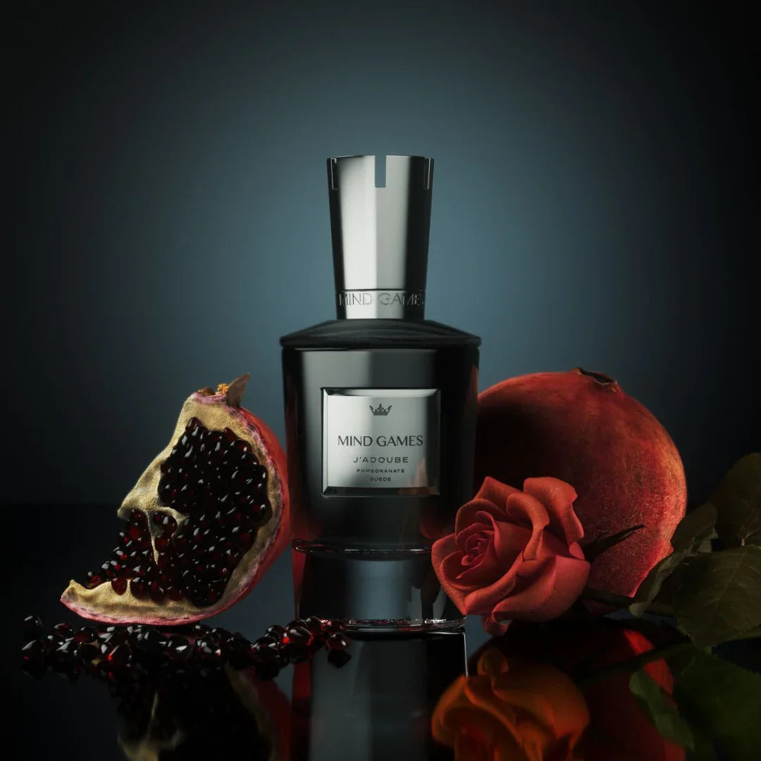 Flakon von "J'Adoube" von Mind Games, umgeben von einer halbierten Granatapfelfrucht, einer Rose und Blättern.