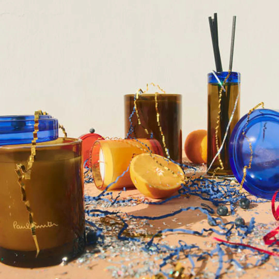Storyteller Duftkerze von Paul Smith, in braunem Glasbehälter mit goldfarbenem Logoschriftzug, umgeben von Dekorationen.