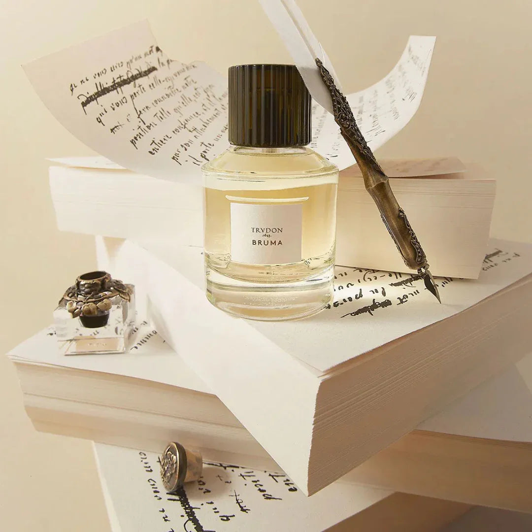Glasflakon "BRUMA" von Trudon, gefüllt mit einer hellgelben Flüssigkeit, umgeben von alten Büchern und Schreibutensilien.