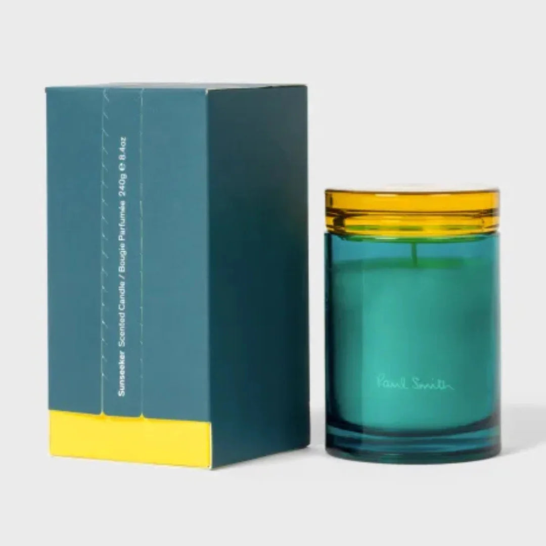 Duftkerze "Sunseeker" von Paul Smith in einem türkisfarbenen Glas mit gelbem Rand, neben der passenden Verpackung.