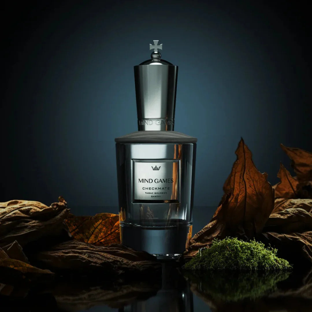 Parfümflakon "Checkmate" von Mind Games, in silbernem und transparentem Design, umgeben von herbstlichen Blättern.