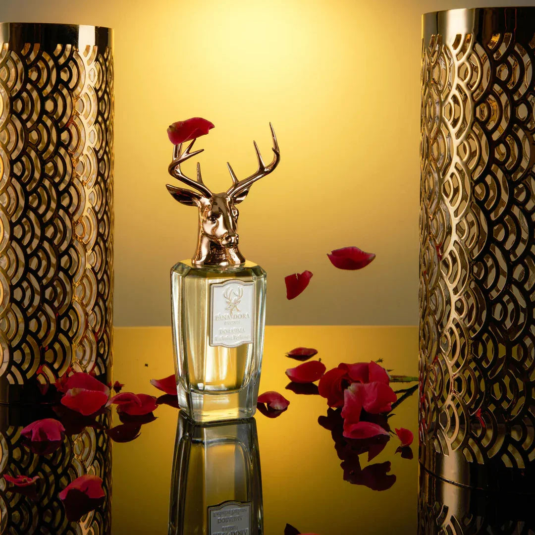 Parfümflakon "Dorasima" von Pana Dora mit Hirschkopf-Design. Umgeben von Rosenblättern und goldenen dekorativen Zylindern.
