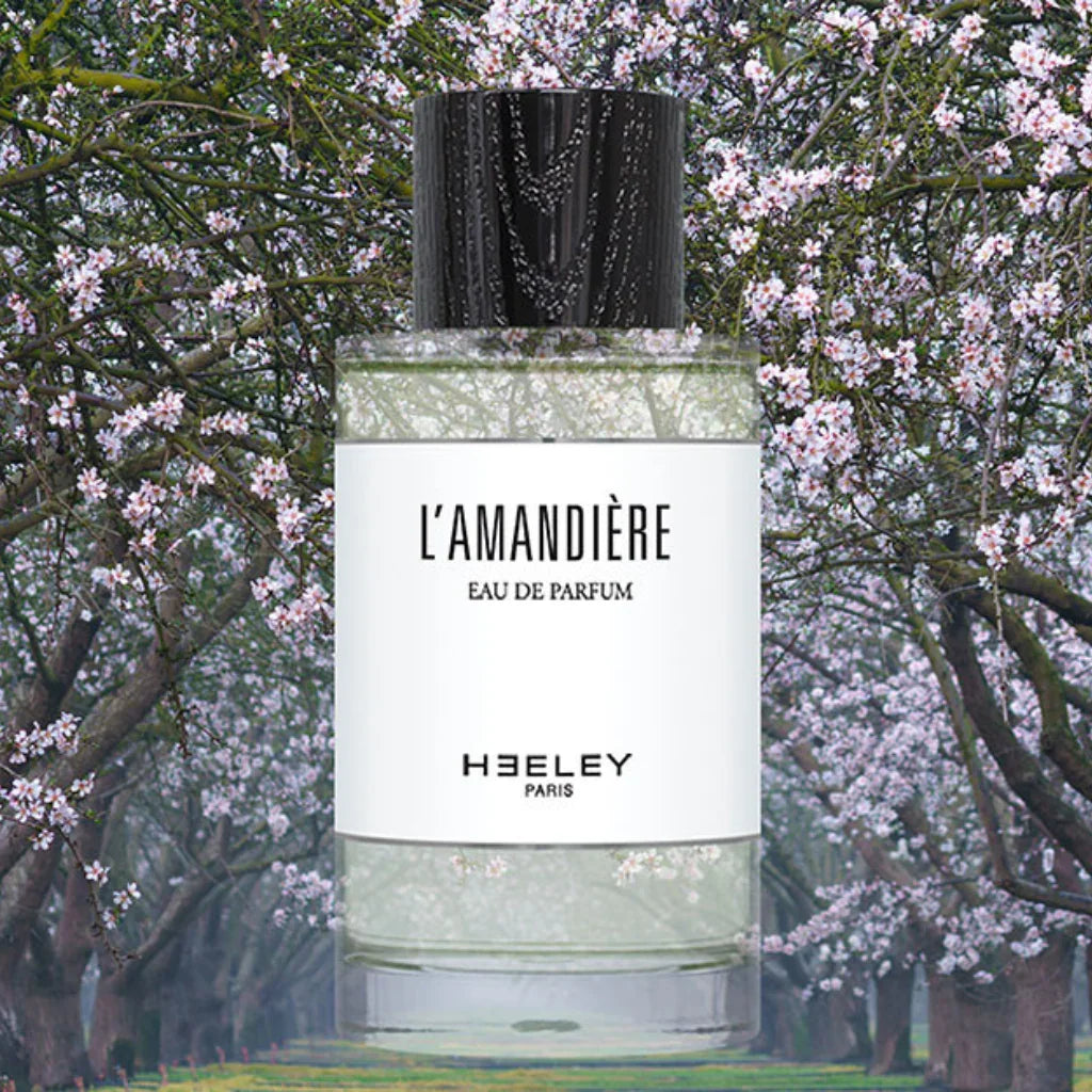 Parfümflasche "L'Amandière" von "Heeley" mit transparentem, zylindrischem Glasbehälter und schwarzem Deckel, umgeben von blühenden Bäumen.