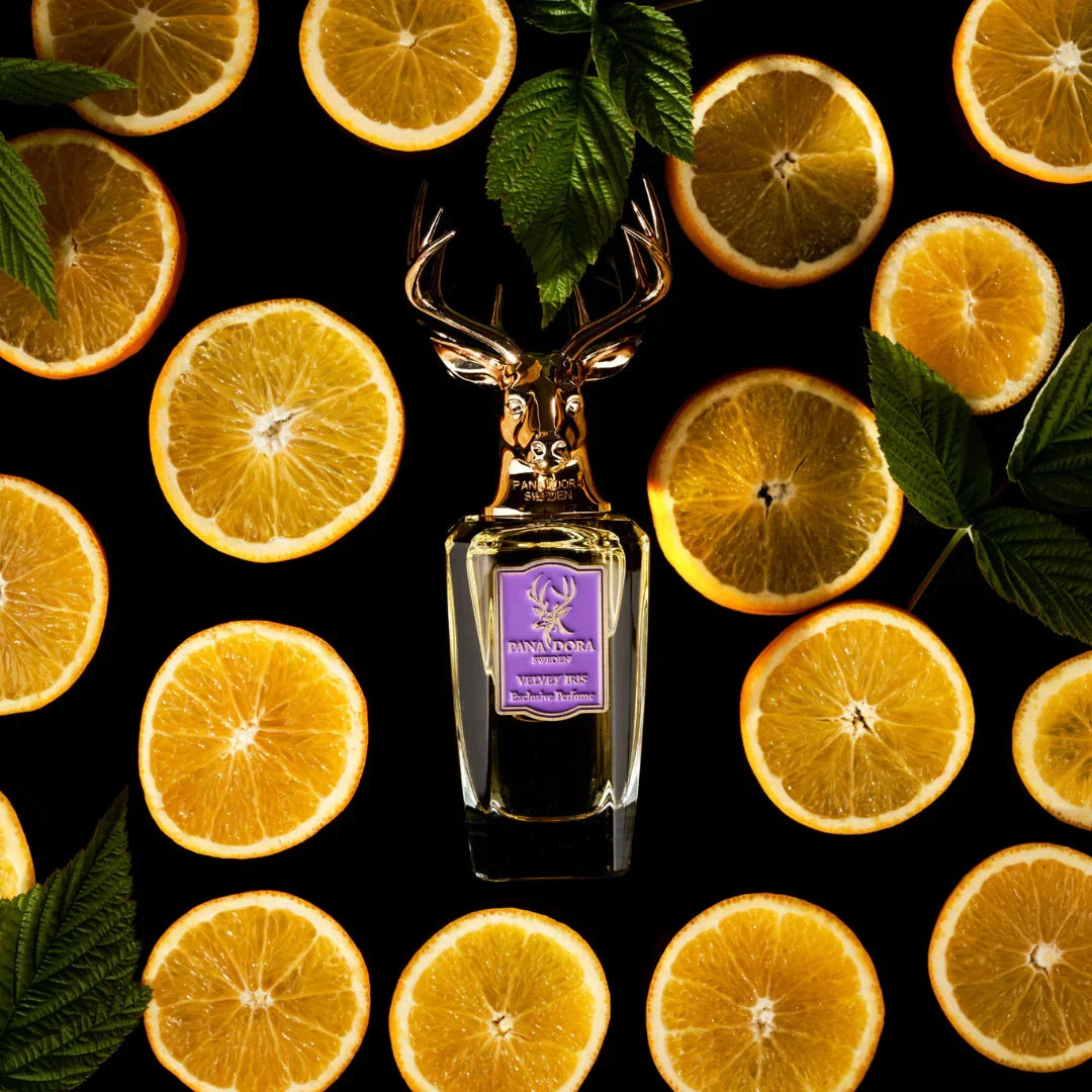 Parfümflasche "Velvet Iris" von Pana Dora, mit goldfarbenem Hirschkopf-Verschluss, umgeben von aufgeschnittenen Orangen und grünen Blättern.