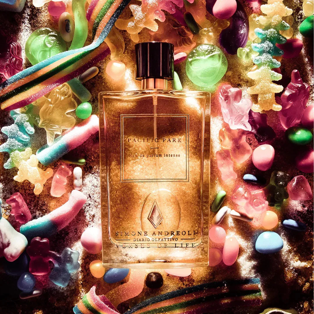 Das Bild zeigt das Parfüm "Pacific Park" von "Simone Andreoli". Die Flasche ist quadratisch und durchsichtig mit schwarzem Verschluss, umgeben von bunten Süßigkeiten.