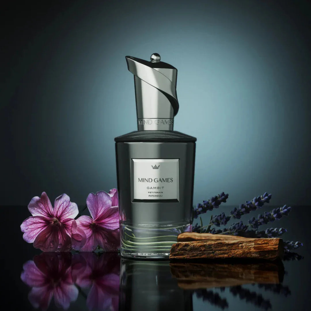 Transparente Parfümflasche "Gambit" von Mind Games mit silbernem Verschluss, umgeben von lila Blumen und einem Holzstück.