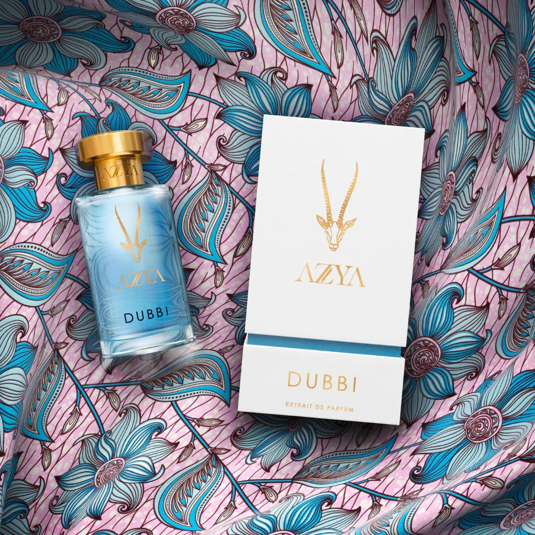 Das Bild zeigt ein Parfümflakon und eine Verpackung der Marke "Azya" mit dem Produktnamen "Dubbi". Der Flakon ist aus Glas mit einem goldenen Deckel und blauer Flüssigkeit. Die Verpackung ist weiß mit goldenen Verzierungen.