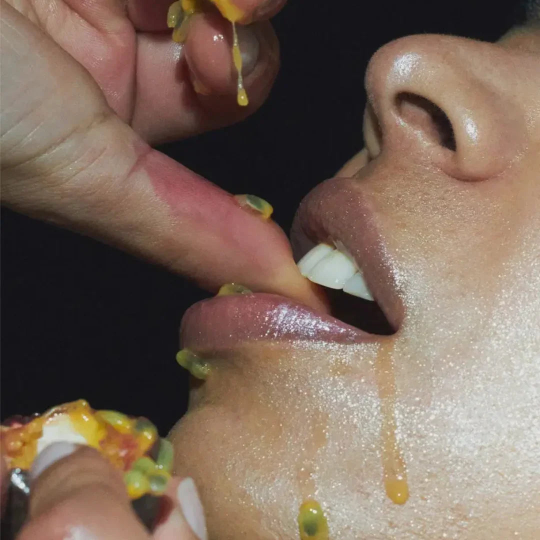 Nahaufnahme eines Gesichts, eine Hand hält eine aufgeschnittene Frucht an den Mund, Saft läuft herunter. Titel: Unfolding Echo, Anbieter: Emil Élise.