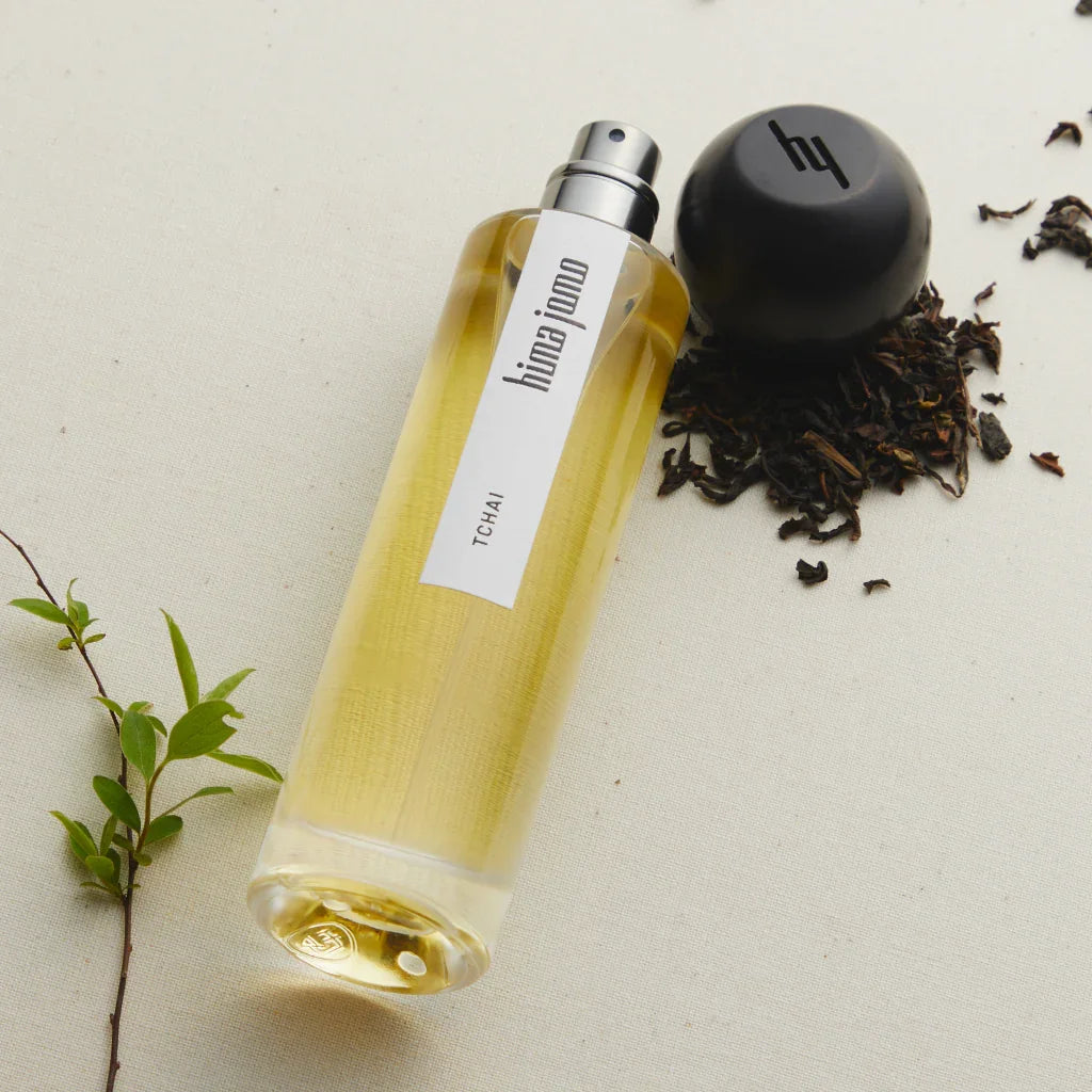 Klarglasflasche mit gelblich-transparenter Flüssigkeit und schwarzem Sprühkopf. Auf dem Etikett steht "hima jomo - Tchai". Daneben liegen Teeblätter und ein schwarzer Deckel mit Logo.