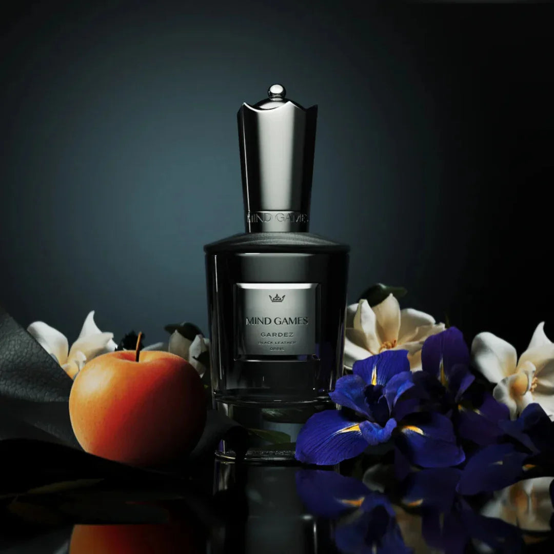 Parfümflakon "Gardez" von Mind Games mit silbernem Verschluss, umgeben von Blumen und einem Apfel.
