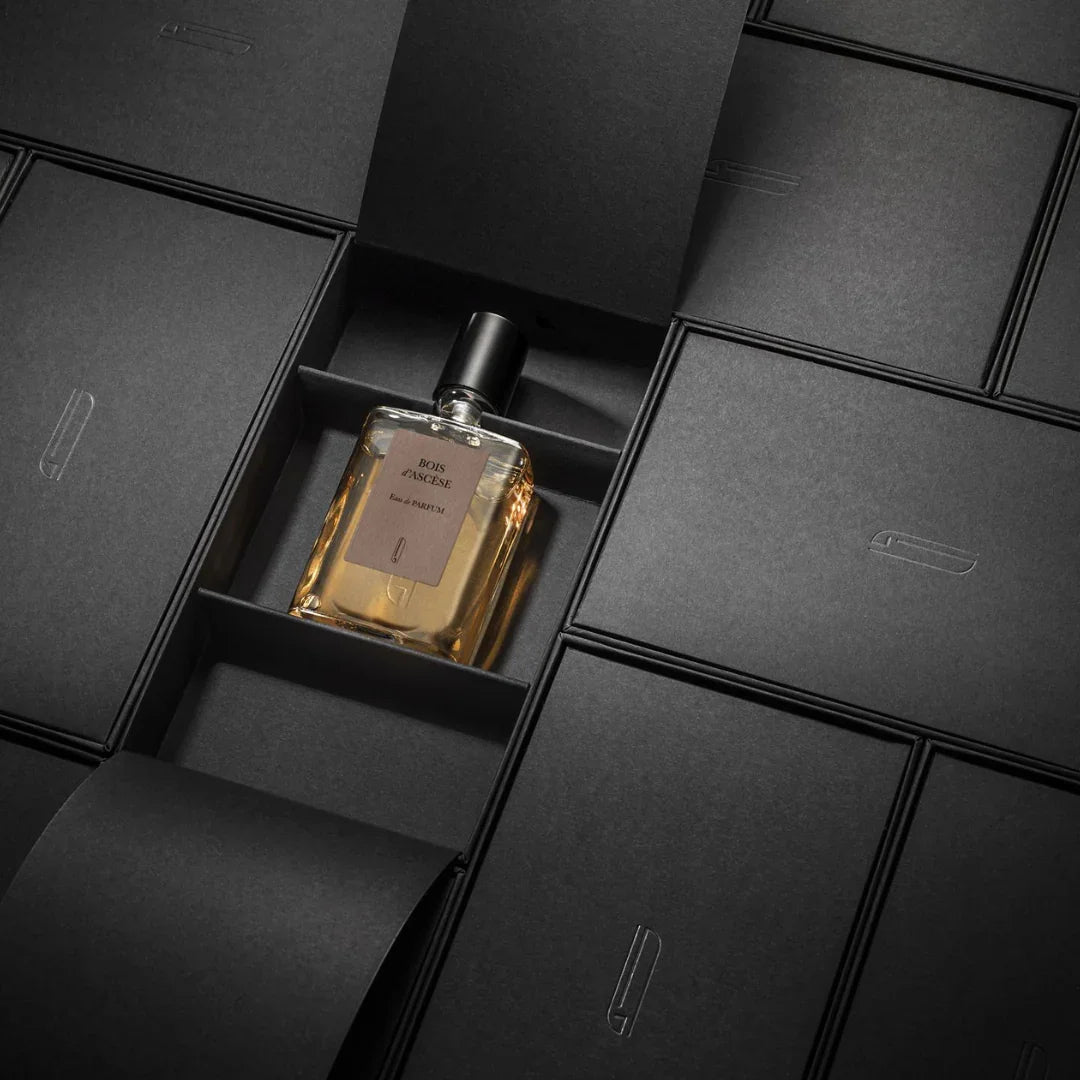 Ein rechteckiger Glasflakon mit schwarzem Verschluss, gefüllt mit einer bernsteinfarbenen Flüssigkeit. Auf dem Etikett steht "Bois d'Ascese" von Naomi Goodsir. Der Flakon liegt in einer schwarzen Verpackung.