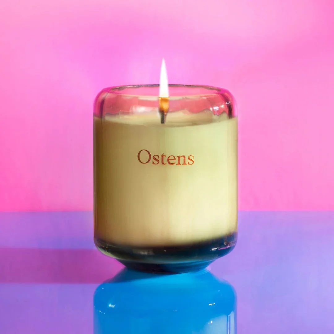 "Illumination Jasmine Duftkerze" von Ostens, eine brennende Kerze in einem transparenten Glas mit "Ostens"-Aufdruck.