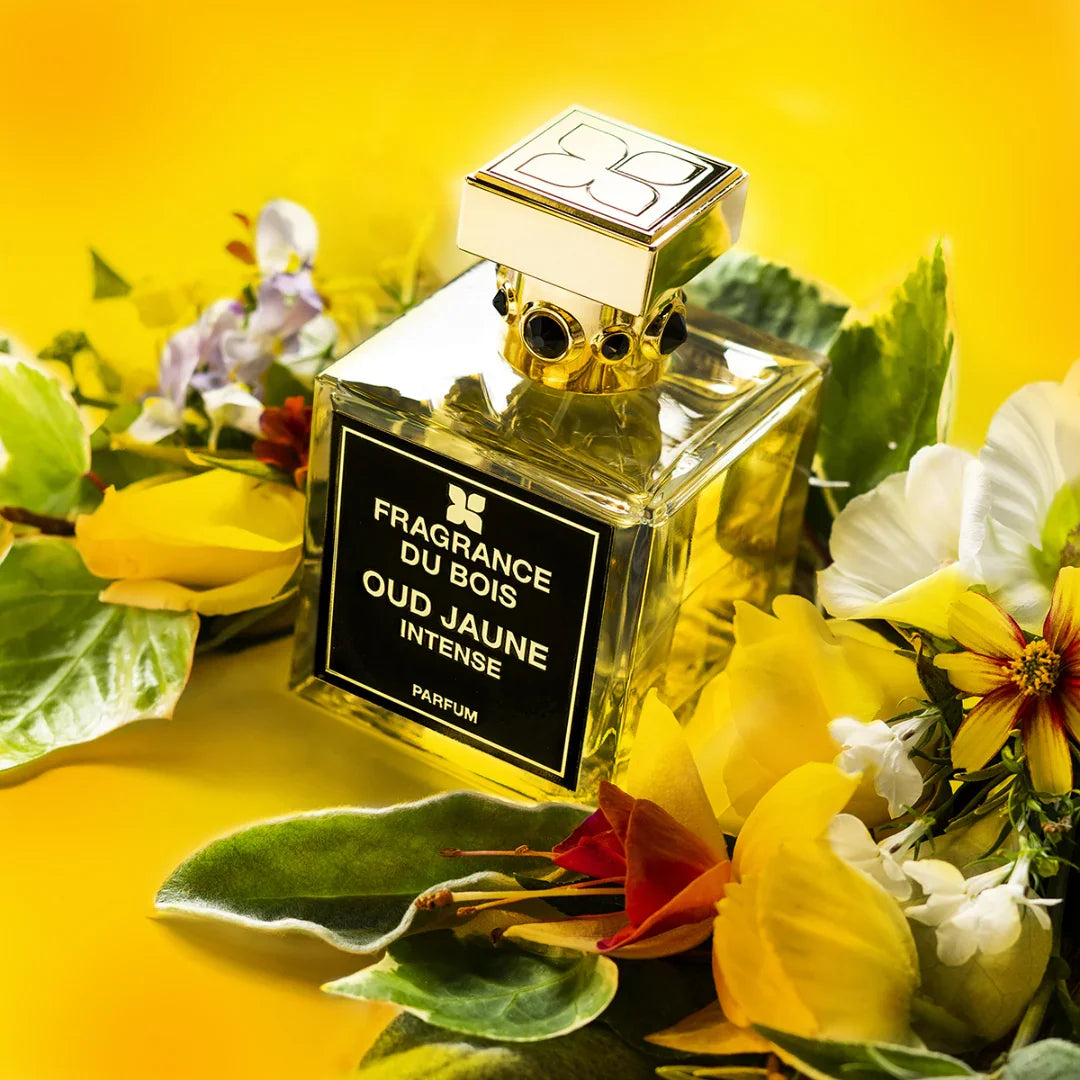 Klares, rechteckiges Glasparfümflakon "Oud Jaune Intense" von FRAGRANCE DU BOIS mit goldener Verzierung, umgeben von Blüten.