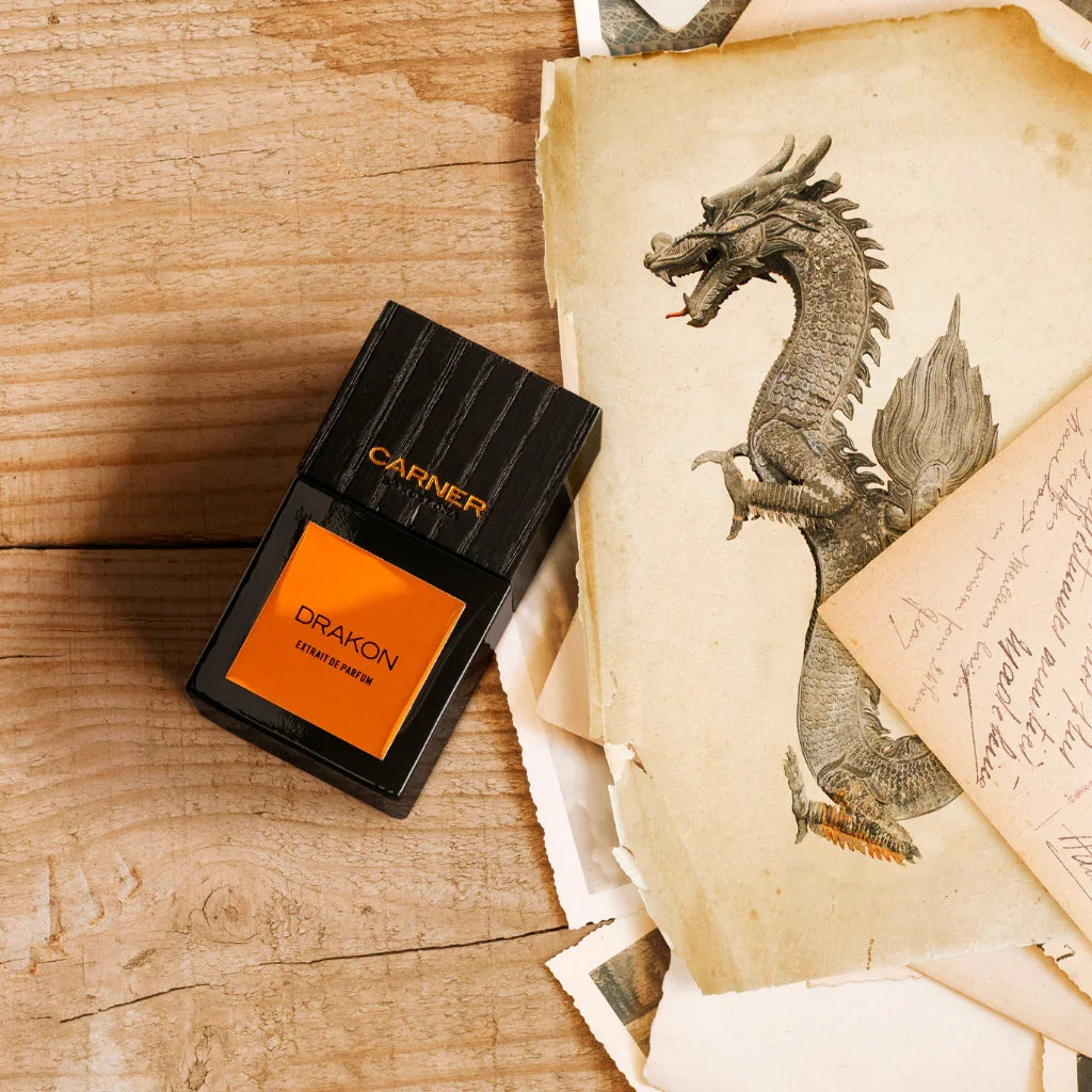 Flasche des Parfums "Drakon" von Carner Barcelona, rechteckig mit schwarzem und orangem Etikett, auf hölzernem Untergrund, neben Zeichnungen eines Drachen.
