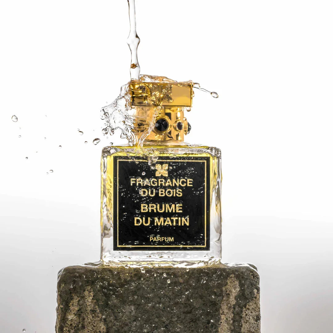 Ein quadratischer Parfümflakon mit goldenem Verschluss, beschriftet mit "FRAGRANCE DU BOIS - BRUME DU MATIN". Flüssigkeit fließt darüber.