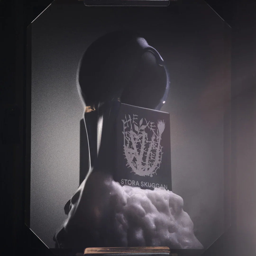 Schwarze, kugelförmige Flasche mit weißem Logo "Hexensalbe" und "Stora Skuggan" auf schwarzem Etikett; sitzt auf schaumartigem Sockel.