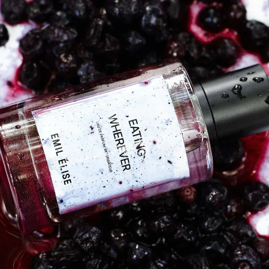 Parfumflasche "Eating Wherever" von Emil Élise, gefüllt mit einer klaren Flüssigkeit, liegt auf dunklen Beeren.