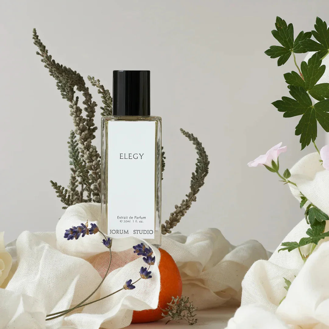 Parfümflakon "Elegy" von Jorum Studio mit schwarzer Kappe und klarer Flüssigkeit, umgeben von Blumen und Stoff.