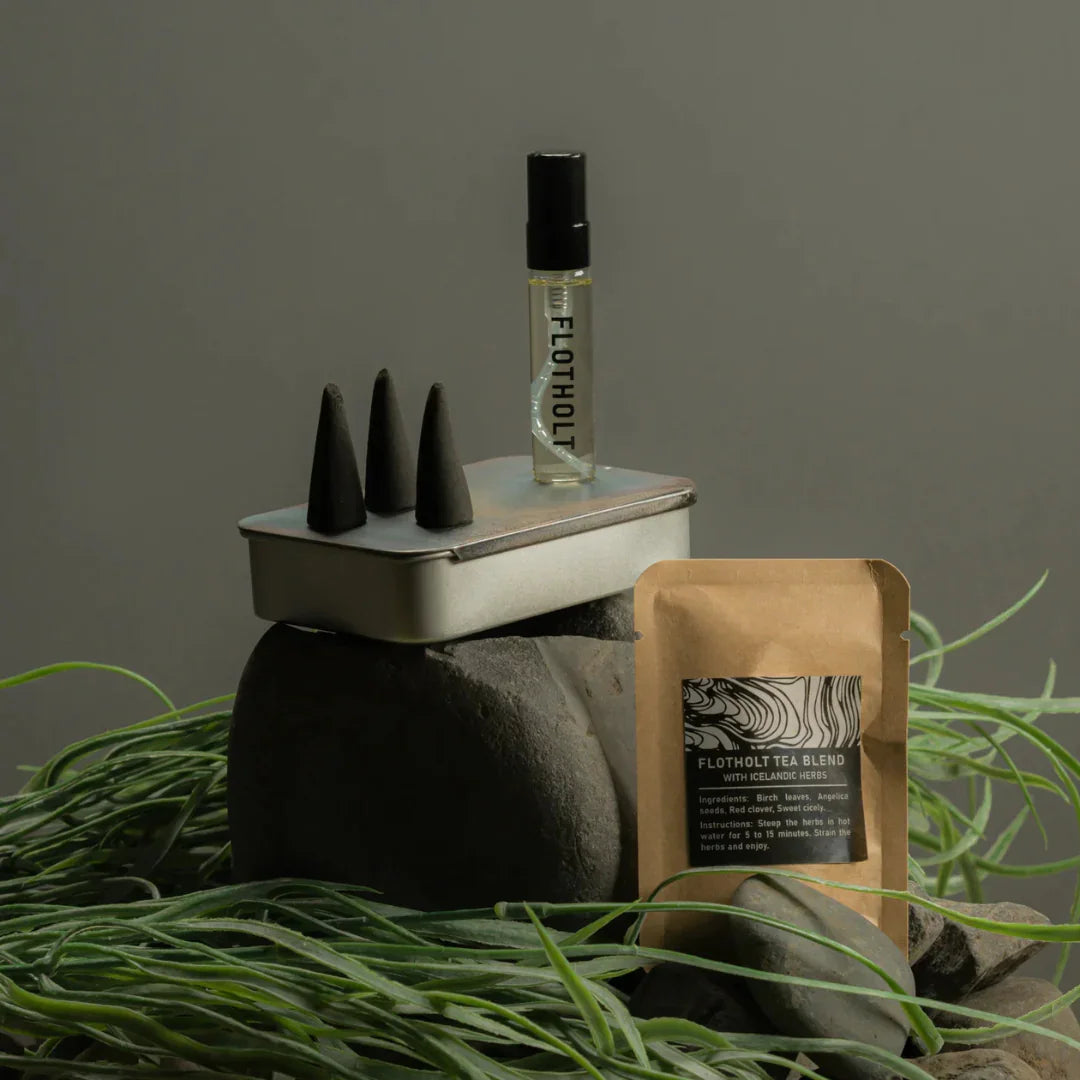 Flotholt Survival Kit von Fischersund zeigt eine Metalldose mit drei schwarzen Kegeln, ein Fläschchen mit "FLOTHOLT"-Aufschrift und eine Teebeutelverpackung auf Steinen und Pflanzen.