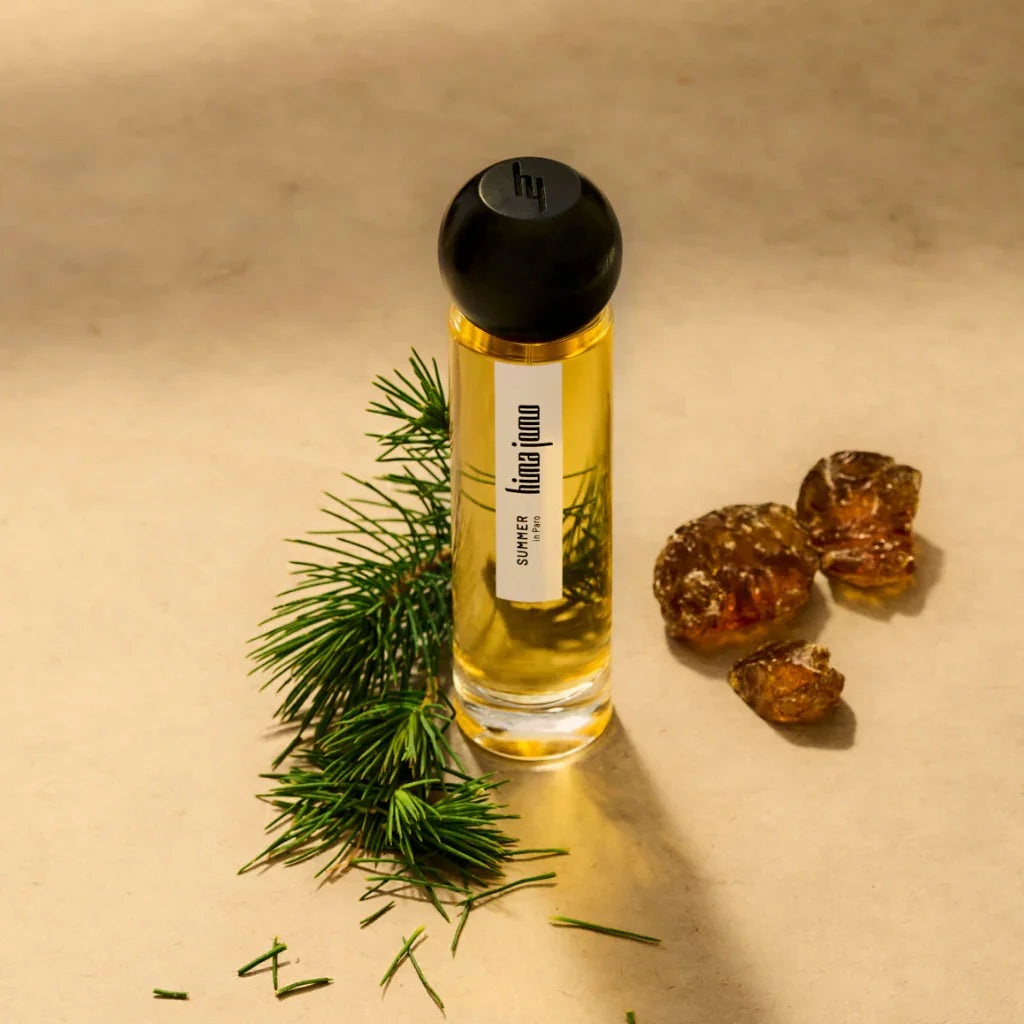 Schmales, zylindrisches Parfümflakon aus Glas mit schwarzem Verschluss und "hima jomo" Etikett; umgeben von Kiefernzweigen und Harzstücken. Produkt: Seasons Collection - Summer on Paro, Hersteller: hima jomo.