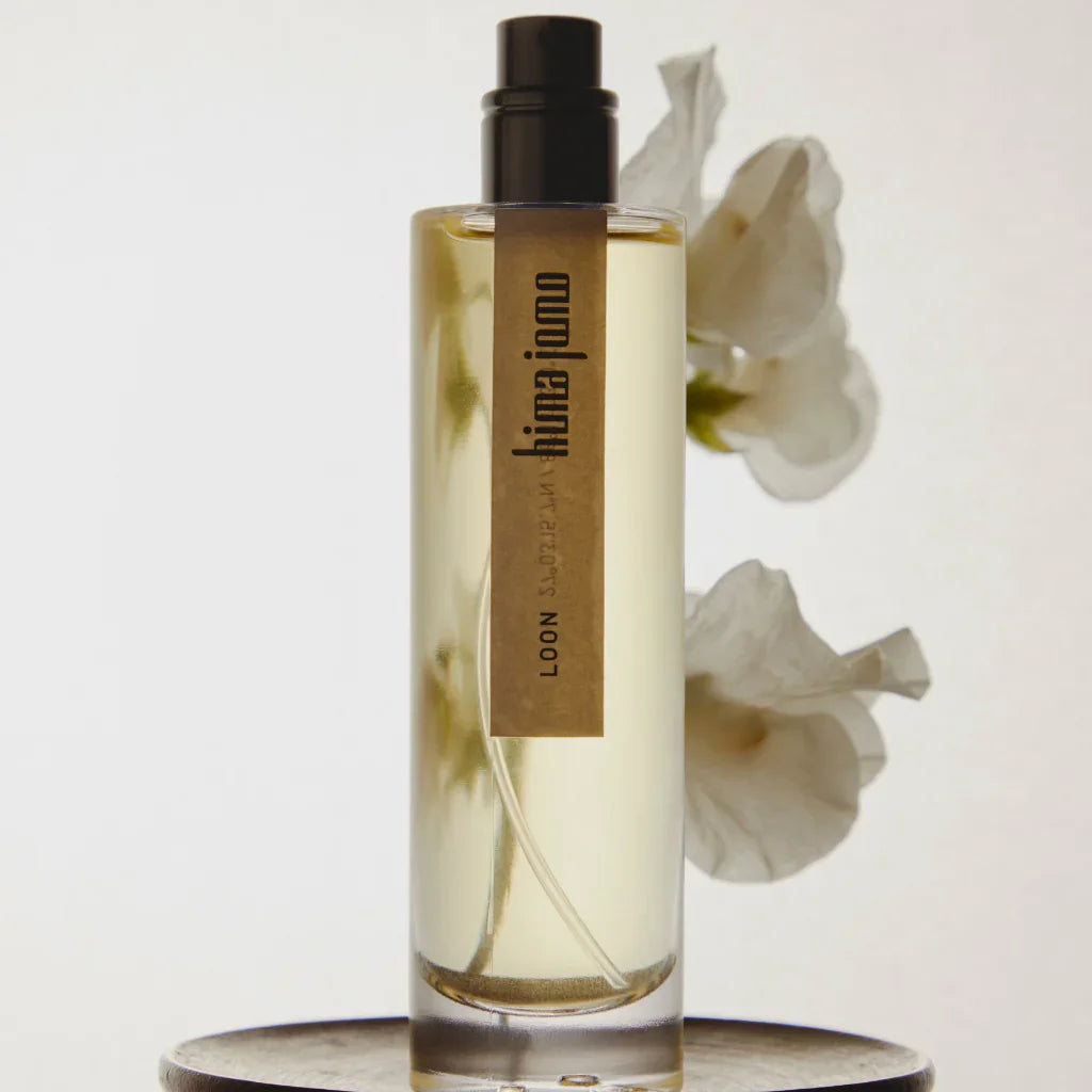 Glasflakon mit hellem Parfüm, schwarzem Sprühkopf und beiger Etikette "hima jomo". Weiße Blüten im Hintergrund.