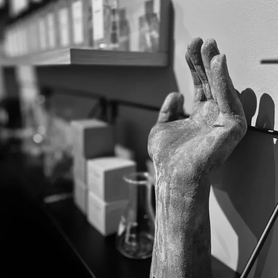 Ein schwarz-weißes Bild zeigt eine skulpturale Hand, gehoben in einer Geste. Im Hintergrund sind Regale mit verschiedenen Flaschen und Schachteln sichtbar. Produkt: Figueres. Anbieter: FUMparFUM.