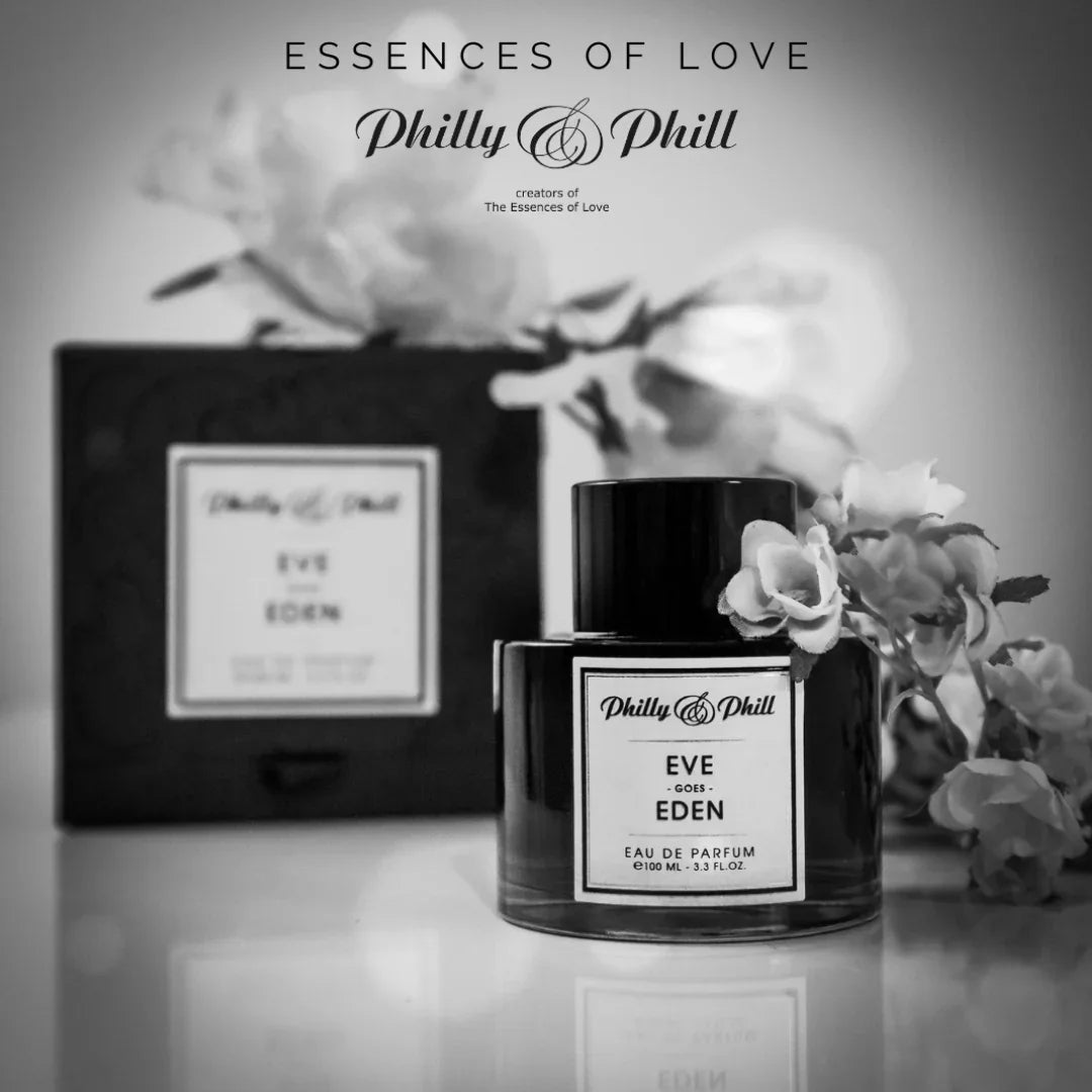Ein schwarzer Parfumflakon mit weißem Etikett "EVE GOES EDEN" von Philly & Phill, umgeben von Blumen, steht vor einer Schachtel.