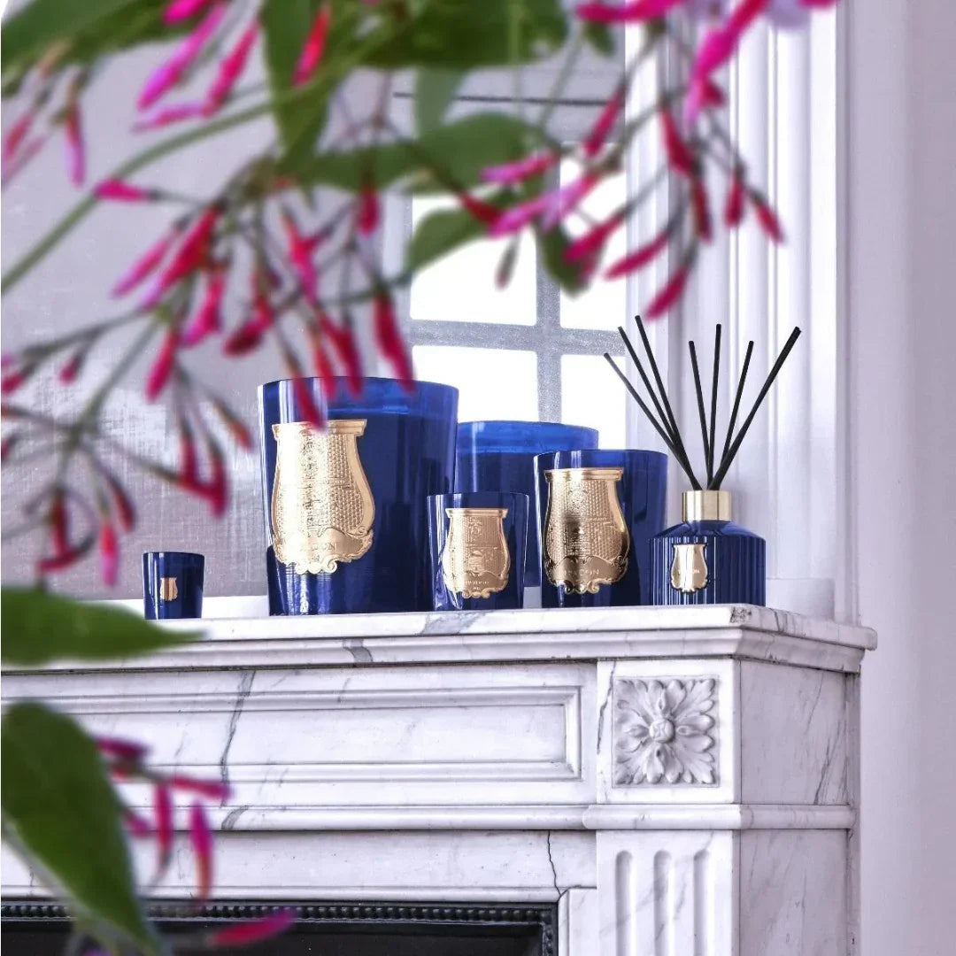 Les Belles Matières - MADURAÏ - Duftkerze von Trudon, mehrere blaue Kerzen mit goldenen Emblemen auf einem Marmorkamin platziert.