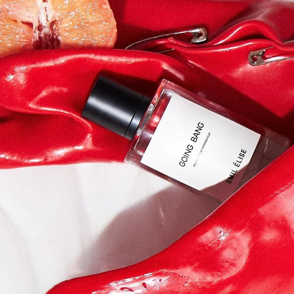 Parfümflasche "Going Bang" von Emil Élise, transparent mit schwarzem Verschluss, liegt zwischen roten glänzenden Stoffen. Grapefruit-Scheibe sichtbar.