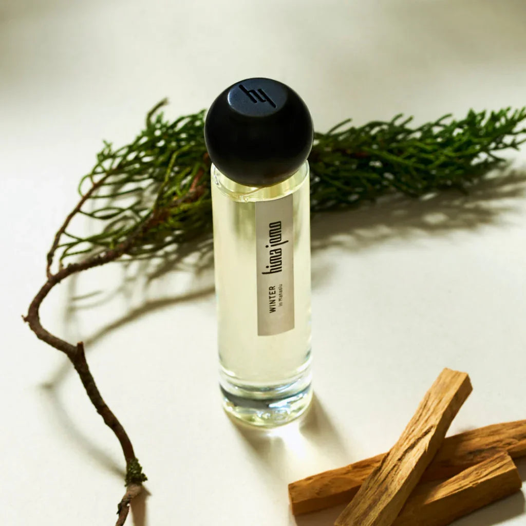 Glasflakon mit durchsichtigem Parfüm, schwarzer Kappe, Label "hima jomo Winter"; umgeben von Zweigen und Holzstücken. Titel: Seasons Collection - Winter in Manaslu. Anbieter: hima jomo.