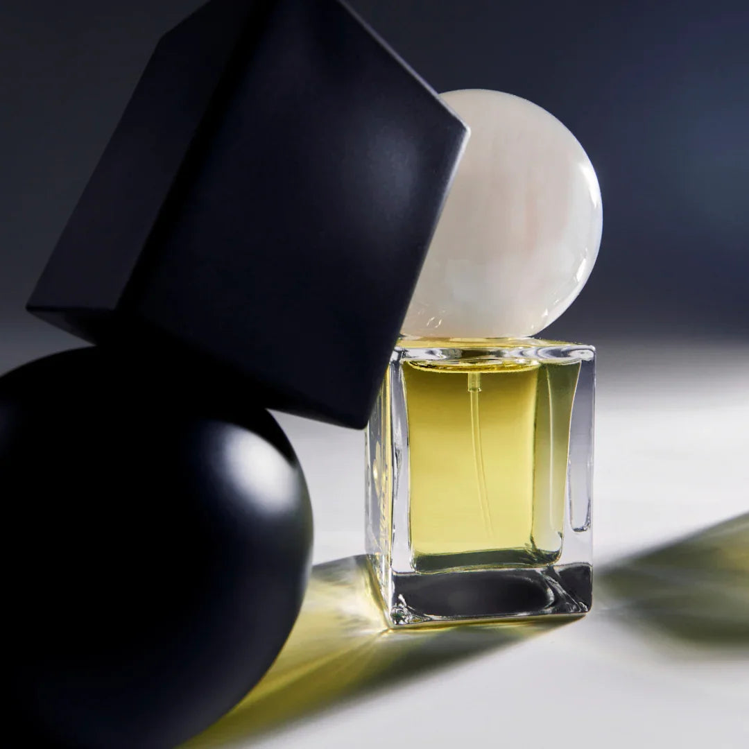 Parfümflakon mit gelblicher Flüssigkeit und transparentem Glas. Große schwarze und weiße geometrische Objekte daneben. Titel: Mistpouffer, Anbieter: Stora Skuggan.