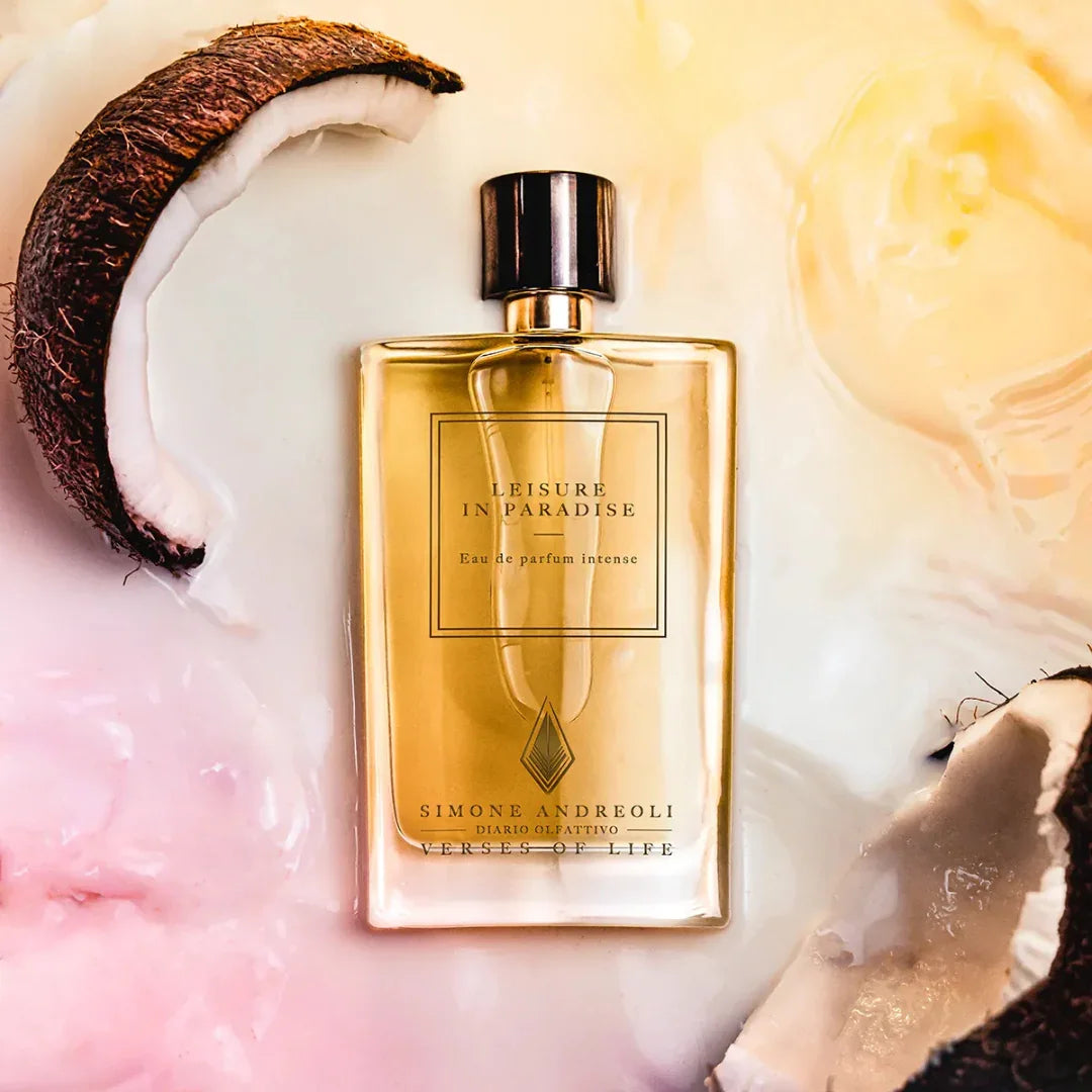 Das Bild zeigt das Parfüm "Leisure in Paradise" von Simone Andreoli. Der Flakon ist rechteckig und durchsichtig, gefüllt mit einer goldenen Flüssigkeit. Der Verschluss ist zylindrisch und metallisch glänzend. Im Hintergrund sind einige aufgebrochene Kokosnusshälften zu sehen.
