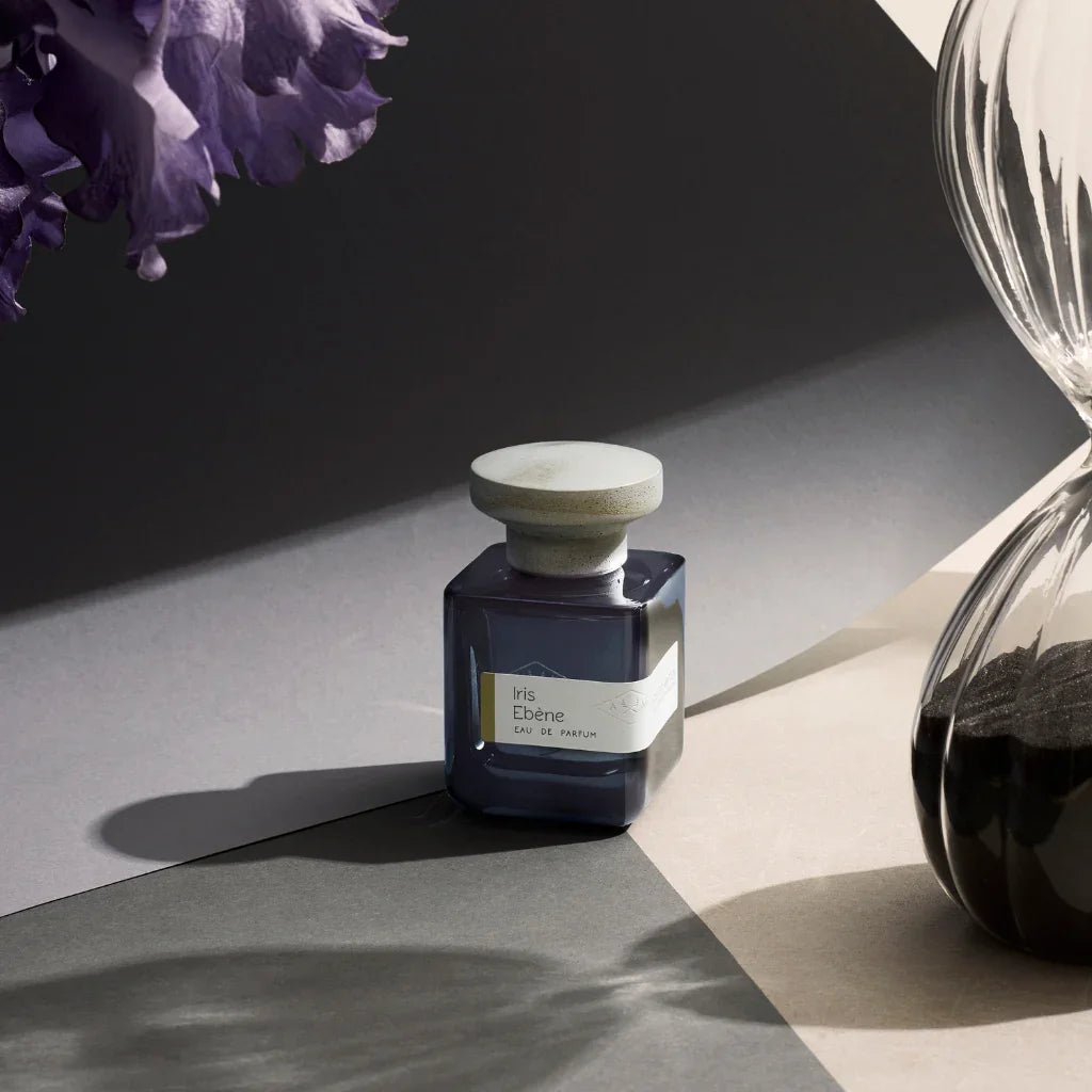 atelier materi iris ebene flakon auf grauem papier neben sanduhr und iris blüte