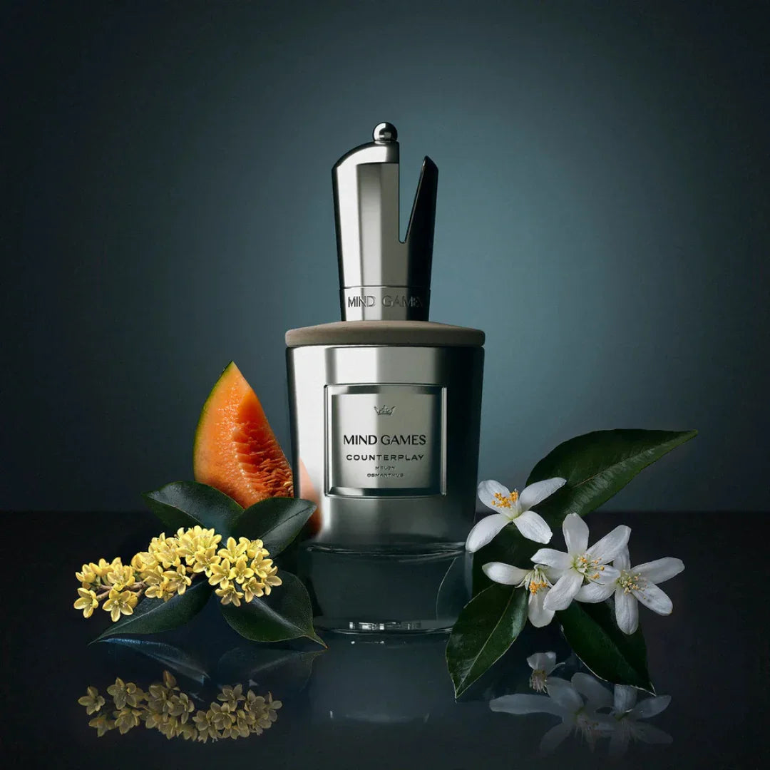 Flakon von Mind Games Counterplay mit glänzendem silbernen Verschluss, umgeben von einer Melone, weißen Blüten und Blättern.