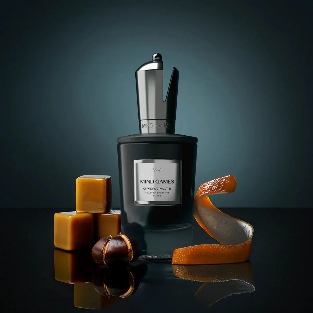 Der Flakon des Eau de Parfum "Opera Mate" von Mind Games steht neben Karamellwürfeln und Orangenschalen.