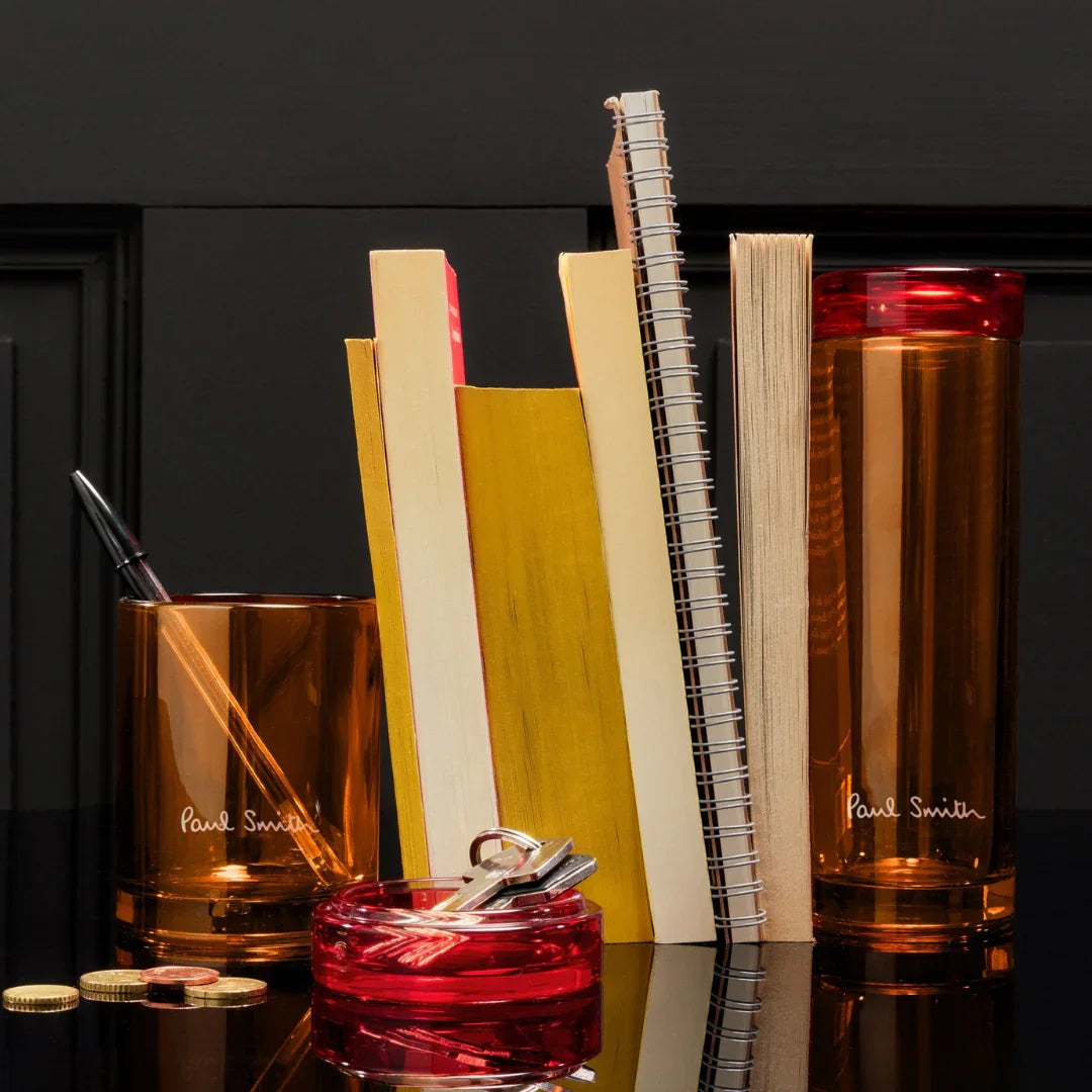 Stilvolle Schreibwaren und Zubehör von Paul Smith, einschließlich Notizbüchern, Stiften und Aufbewahrungsbehältern in warmen Farbtönen. Produkt: Bookworm Duftkerze.