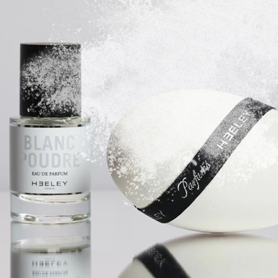 Parfümflasche "Blanc Poudre" von Heeley, transparent mit weißem Etikett und schwarzem Deckel, neben einem weißen ovalen Objekt mit schwarzem Band.